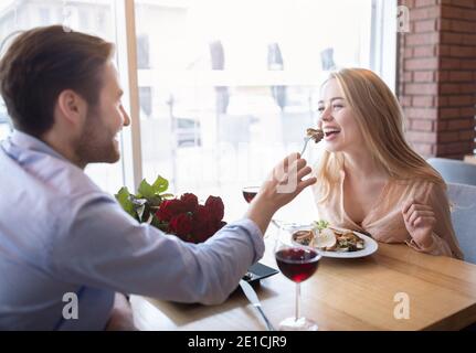 Schönes junges Paar mit romantischen Abendessen mit Blumen und Wein im Café, Millennial Kerl Fütterung seiner Freundin Stockfoto