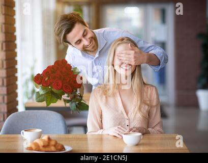 Glücklicher Millennial Kerl, der romantische Überraschung für seine geliebte Frau macht, ihr Blumen gibt und ihre Augen im Café bedeckt Stockfoto