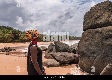 Afrikanische Frau steht an einem schönen Strand mit dunklen großen Steinen und sauberen Strand am Rande des Dschungels in Axim Ghana Westafrika Stockfoto