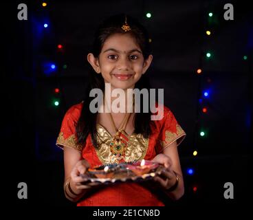 Ein schönes lächelndes indisches Mädchen in ethnischer Kleidung mit gekleidet Juwelen und halten Öllampen feiern diwali in der Nacht mit Dunkles Licht im Hintergrund Stockfoto