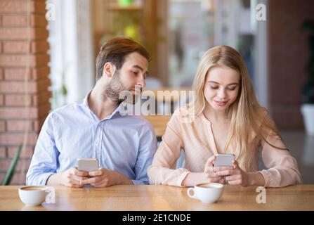 Millennial Paar mit stumpfen Datum, stecken in Smartphones, eifersüchtigen jungen Mann Blick in Frau Gadget im Café Stockfoto