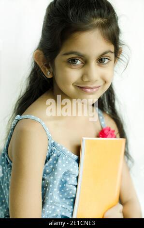 EIN WUNDERSCHÖNES PORTRÄT EINES KLEINEN LÄCHELNDEN INDISCHEN SCHULMÄDCHEN, DAS EIN BUCH MIT DER DARSTELLUNG DES LERNENS ZU HAUSE WÄHREND DER COVID-19 HAUSQUARANTÄNE HÄLT. Stockfoto