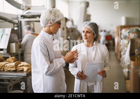 Fokussierte engagierte weibliche Mitarbeiterin in sterilen Kleidung reden über Fortschritte in der Lebensmittelfabrik . Stockfoto