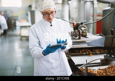 Junge Arbeiterin in steriler Kleidung überprüft die Qualität der Produkte in der Lebensmittelfabrik. Stockfoto