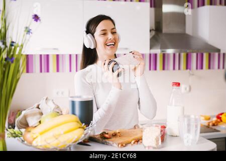 Fröhliche und schöne junge Frau schüttelt ihre Schüttelschale Gefüllt mit Smoothie Zutaten, während jamming zu Musik auf sie Kopfhörer in der hellen Stockfoto
