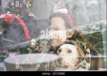 Blick durch das Fenster der jungen Frau neben Weihnachtsbaum mit ihren Hunden mit einem von ihnen küssen sie sitzen, mit verschneiten Natur reflektiert in der Stockfoto