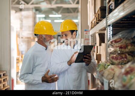 Zwei Kollegen sprechen und lächeln, während sie im Lager stehen. Jüngere mit Tablet. Auf Köpfe Helme. Stockfoto