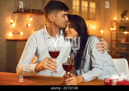 Liebevoller Ehemann umarmt seine Frau zärtlich und küsst ihre Stirn, während er in der Küche sitzt. Stockfoto