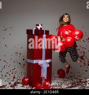 Schöne kaukasische Frau freut sich und springt neben roten Boxen mit Geschenke mit vielen roten Ballonen mit Verkauf und Prozentsätze auf sie Stockfoto