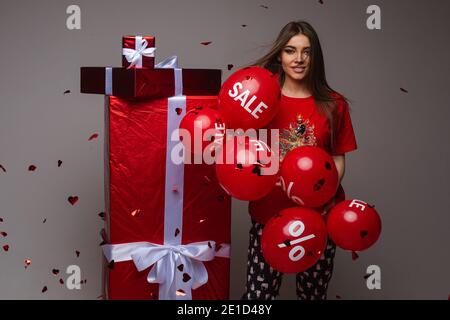 Schöne kaukasische Frau steht in der Nähe von roten Boxen mit Geschenken mit Eine Menge roter Ballone mit Umsatz und Prozentsätzen auf Sie Stockfoto