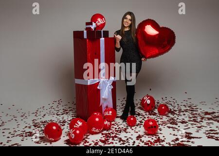 Schöne kaukasische Frau posiert für die Kamera in der Nähe von roten Boxen Mit Geschenken und vielen roten Ballonen mit Verkaufszahlen Und Prozent auf sie Stockfoto