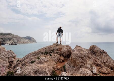 Der junge Mann steht auf einem Felshügel mitten im mittelmeer. Blick auf Meer und Klippen in der Ferne. Berühmter und schöner Ort in Zypern Stockfoto