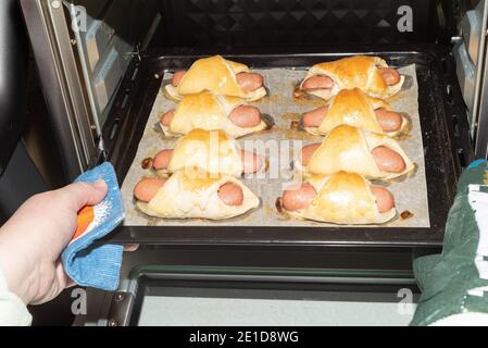 Die Frau nimmt aus dem Ofen ein heisses Backblech mit vorbereiteten Würstchen in den Teig. Kochen im Ofen. Stockfoto