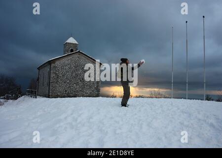 Mädchen im Winter Overalls spielt mit weichem weißen Schnee in Eine wunderschöne Sonnenuntergangslandschaft am Meer unter einer Kirche Mit Kirchturm in den Bergen in apuan al Stockfoto