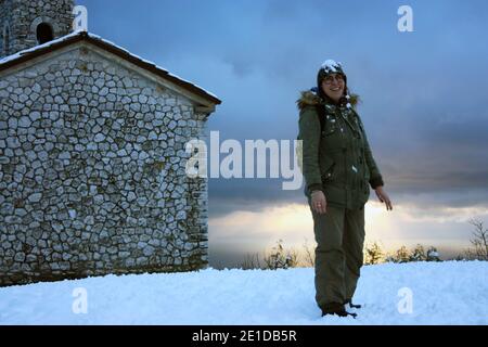 Mädchen im Winter Overalls spielt mit weichem weißen Schnee in Eine wunderschöne Sonnenuntergangslandschaft am Meer unter einer Kirche Mit Kirchturm in den Bergen in apuan al Stockfoto