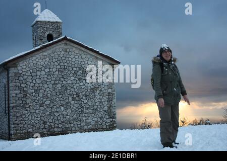 Mädchen im Winter Overalls spielt mit weichem weißen Schnee in Eine wunderschöne Sonnenuntergangslandschaft am Meer unter einer Kirche Mit Kirchturm in den Bergen in apuan al Stockfoto