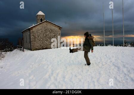 Mädchen im Winter Overalls spielt mit weichem weißen Schnee in Eine wunderschöne Sonnenuntergangslandschaft am Meer unter einer Kirche Mit Kirchturm in den Bergen in apuan al Stockfoto