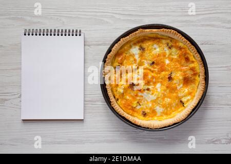 Hausgemachtes Bacon Quiche mit Eiern und Cheddar-Käse, leerer Notizblock auf einer weißen Holzoberfläche, Blick von oben. Flach liegend, von oben, Draufsicht. Speicherplatz kopieren Stockfoto