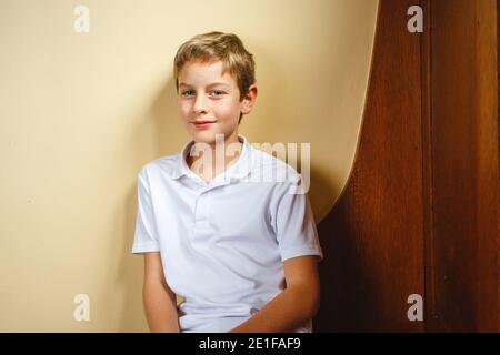 Ein kleiner Junge sitzt auf einer Holzbank und lehnt sich an die Wand Mit direktem Blick Stockfoto