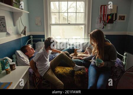 Drei Geschwister sitzen auf einem Bett kuscheln mit einem niedlichen Familienhund im Schlafzimmer