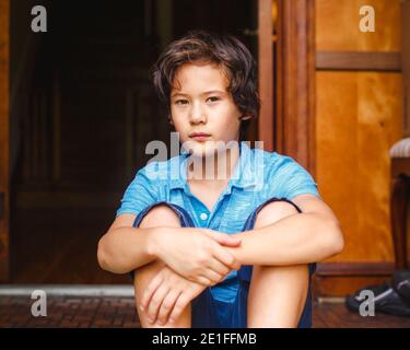 Ein Junge mit goldener Haut und ernstem Ausdruck sitzt darin Eingang des Hauses Stockfoto