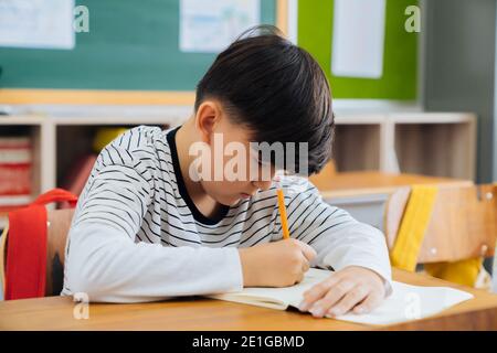 Kaukasischer Schuljunge sitzt in der Schule mit Bleistift, studieren, Bildung, Lernen. Männlicher Student sitzt am Schreibtisch im Klassenzimmer schriftlich in Notebook in Prüfung. Stockfoto