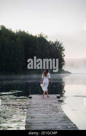 Junge Frau in weißem Kleid auf einem See an einem kühlen Morgen mit einem Nebel über Wasser. Stockfoto