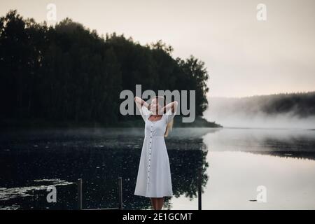 Junge Frau atmet tief, während sie auf einem hölzernen Pier steht Bei Sonnenaufgang am nebligen See Stockfoto