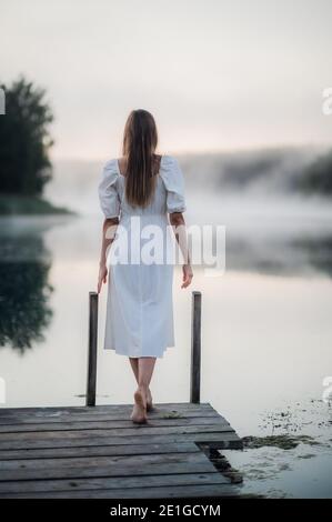 Rückansicht einer jungen Frau in weißem Kleid, die allein auf dem Steg steht und auf den See starrt. Neblig kühl Morgen mit einem Nebel über Wasser. Stockfoto