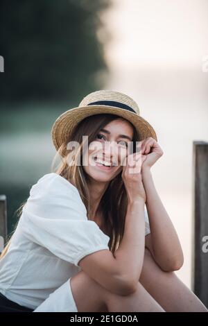 Porträt einer glücklichen kaukasischen Frau in einem weißen Kleid und Hut im Freien sitzen und lächeln. Stockfoto