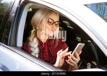 Eine blonde Frau in einer Brille in einer roten Bluse sitzt hinter dem Steuer und löst geschäftliche Angelegenheiten mit dem Telefon. Beschäftigung. Stockfoto