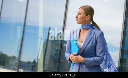 Eine junge blonde Frau in einem formellen Anzug hält Dokumente auf dem Hintergrund eines Geschäftsgebäudes. Arbeitskonzept. Stockfoto