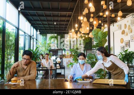 Asiatische Frau waering Maske und asiatische Menschen Essen allein am Tisch mit Plexi Bildschirm zwischen Tisch in Wiedereröffnung Restaurant nach Lockdown Maßnahme. Stockfoto