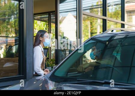 Asiatischer Mann in Schutzmaske bestellen Essen und Kaffee mit Frau Kellnerin tragen Gesichtsmaske und Gesichtsschutz am Drive-Thru während Coronavirus Ausbruch. Stockfoto