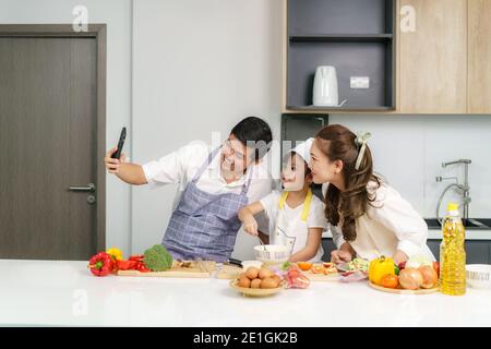 Junge asiatische Familie bereiten Salat in der Küche und Vater nehmen ein Foto Selfie per Telefon. Aufgeregt lächeln und Fällen glücklich. Eltern lehren Tochter Stockfoto