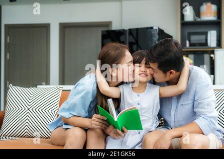 Glückliche asiatische Familie mit Vater und Mutter küssen in ihre Tochter Wange zusammen, während im Wohnzimmer zu Hause sitzen. Liebe Emotion, lächelndes Gesicht, enjo Stockfoto
