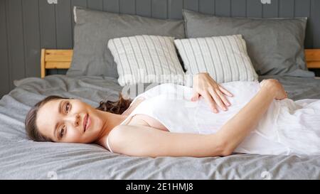 Hübsche junge schwangere Frau in weißem Kleid liegt auf Doppel Bett mit grauer Bettwäsche und Designer-Kissen, die den Bauch streicheln Lächelnd im Zimmer aus der Nähe Stockfoto