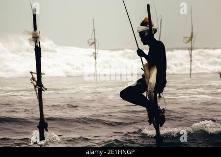 Weligama, Sri Lanka - 07 26 2020: Alte Fischer hält einen Ritt in einem Holzpfahl während sitzen, Angeln in der Abendsilhouette, Ozeanfluten krachen Stockfoto