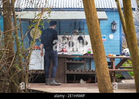 Mann kocht mit einer Grundausstattung verbunden mit umgebauten Zug Wagen auf einem Campingplatz in Fairford, Gloucestershire, England Stockfoto