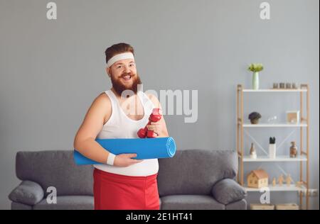 Lustige rote dicke Mann mit einer Matte und Hanteln in den Händen lächeln, während zu Hause stehen. Stockfoto
