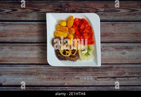 Rindersteak mit Zwiebelringen, Kroketten und Gemüse (Fenchel und Paprika) auf einem weißen quadratischen Teller mit dunklem Holzhintergrund. Deutsche Hausmannskost. Stockfoto