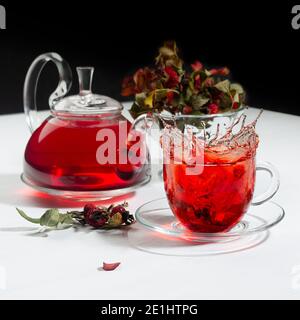 Herbstabend Teezeit mit Spritzer in roter Kräuterteetasse, transparente Teekanne mit Weißdornbeeren, Hagebutte, trockene Blätter auf weißem Holztisch, schwarz w Stockfoto