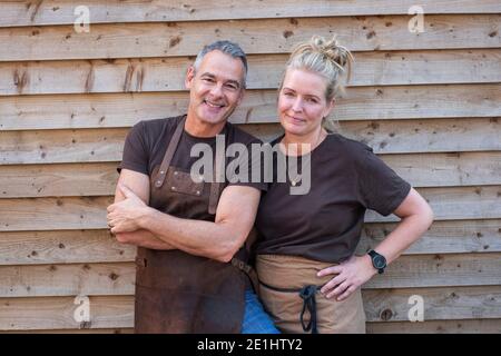 Kellner mit kurzen grauen Haaren und Kellnerin mit blonden Haaren, beide tragen braune Schürzen, lehnen sich an die Holzwand und lächeln an der Kamera. Stockfoto