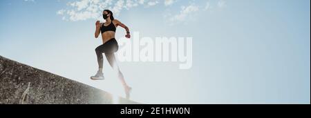 Low-Angle-Aufnahme einer Frau beim Laufen im Freien mit Sonne scheint von hinten. Weiblich in Sportbekleidung und schützende Gesichtsmaske Ausübung Outdoo Stockfoto
