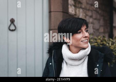 Fröhliche Frau mit kurzen Haaren trägt Winterkleidung und lächelt Vor ihrem Haus Stockfoto