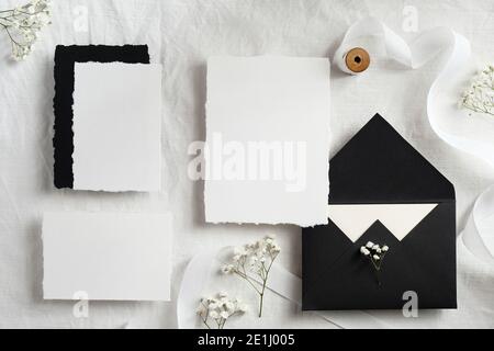 Elegantes Set von Hochzeitspapier Draufsicht. Flach legen leere Papierkarten, schwarze Umschläge, Blumen, Band auf weißem Textilhintergrund. Stockfoto