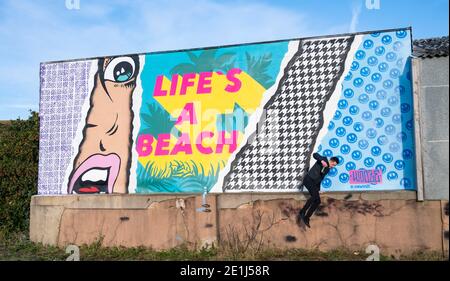 Lancing West Sussex UK 7. Januar 2021 - der Street-Artist Luke Henley, bekannt als Wooze, setzt seinen letzten Schliff an seinem neuesten Stück "Life's A Beach" an der Lancing Seafront in der Nähe von Worthing in West Sussex . Der Künstler erhielt die Erlaubnis, das Kunstwerk auf einer Wand hinter einer lokalen Garage mit Blick auf Lancing Green zu schaffen und er hofft, dass es die Gegend für Wanderer und Besucher erhellt : Credit Simon Dack / Alamy Live News Stockfoto