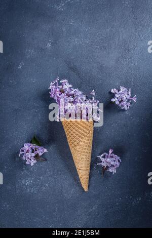 Waffelkegel mit lila Flieder auf dunkelblauem Stein Beton Tischhintergrund. Morher Day Holiday Konzept Stockfoto