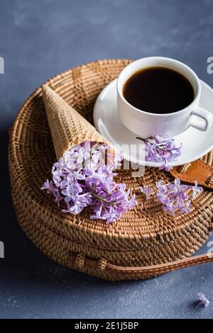 Modische Rattan-Tasche, Tasse Kaffee und lila Flieder in Waffelkegel auf blauem Beton Hintergrund. Konzept Der Spring Summer Card. Stockfoto