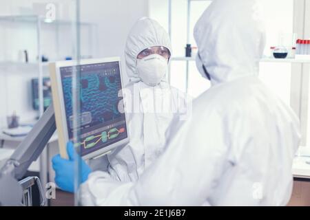 Coworkers im Overall diskutieren über die Evolution von Viren in PSA-Anzug in modernen Gesundheitseinrichtungen gekleidet. Ärzte untersuchen die Evolution des Virus mit Hilfe von High-Tech-Forschung Diagnose gegen covid19 Stockfoto